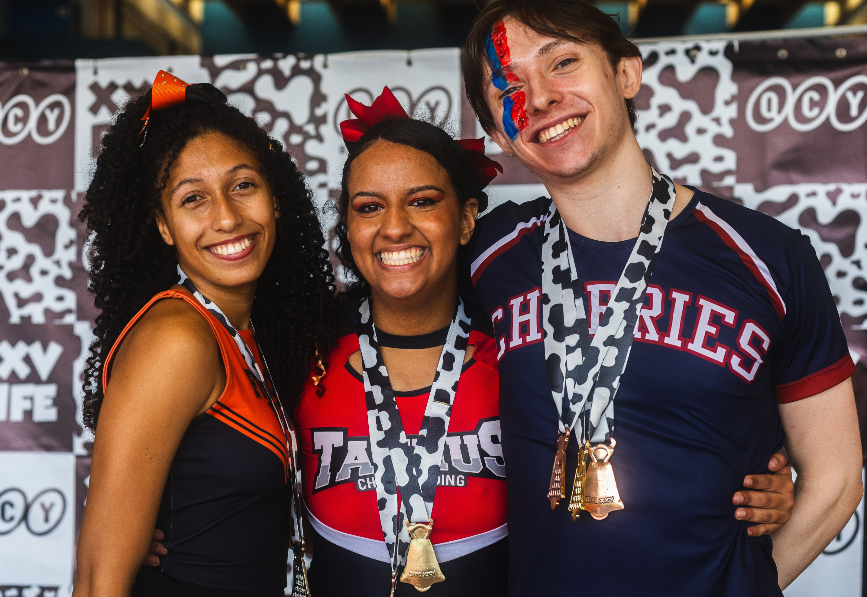 Drei lächelnde Cheerleader mit Medaillen vor QCY-Banner feiern den Erfolg bei den BIFE Games.