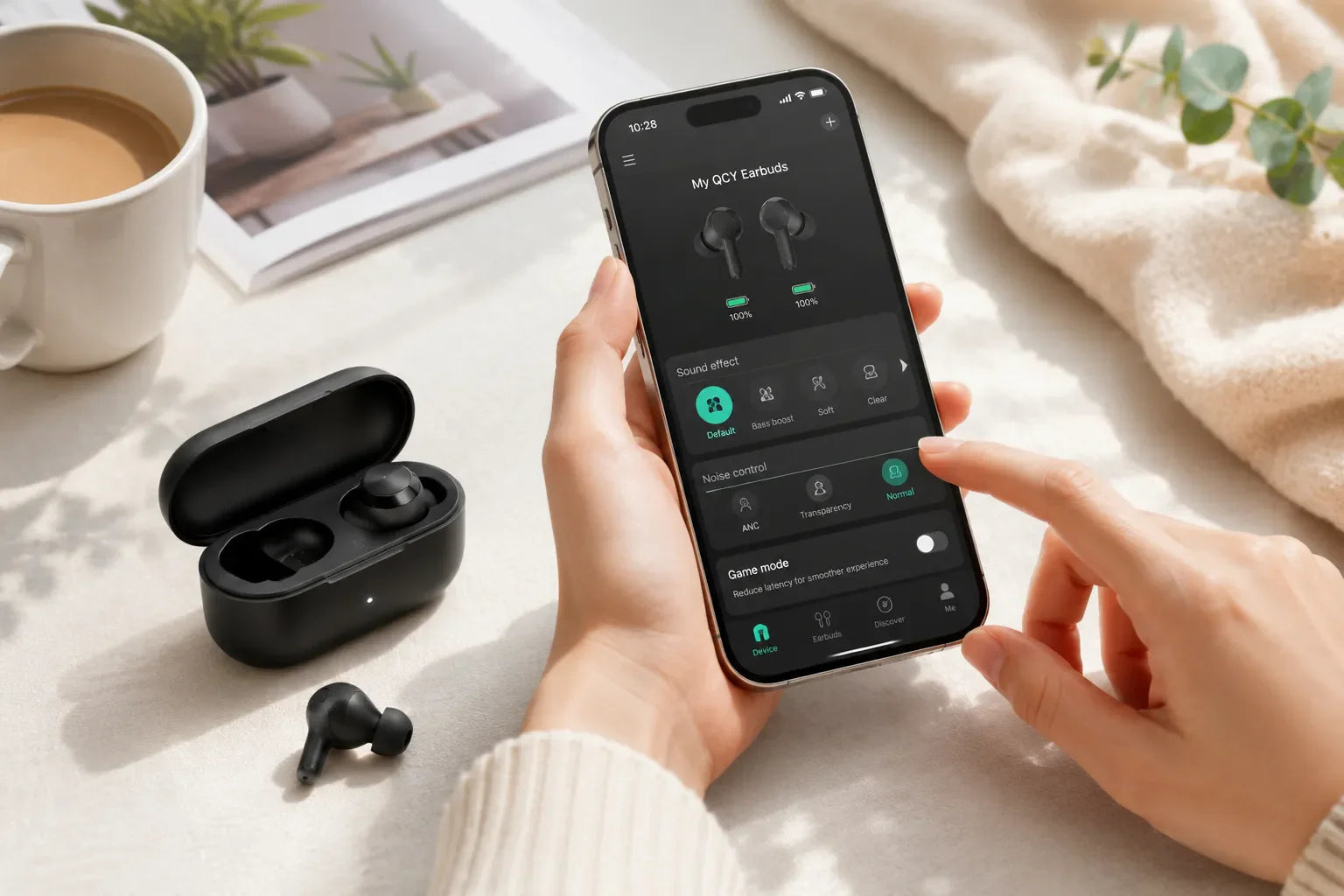 Person bedient QCY App auf Smartphone, um In-Ear-Kopfhörer mit ANC & Soundeffekten zu steuern – neben Ladecase und Kaffee.